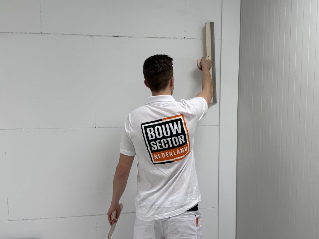 Stukadoor van Bouwsector Nederland zet een lange spaan hoog tegen de wand, met een kleine spaan in zijn andere hand