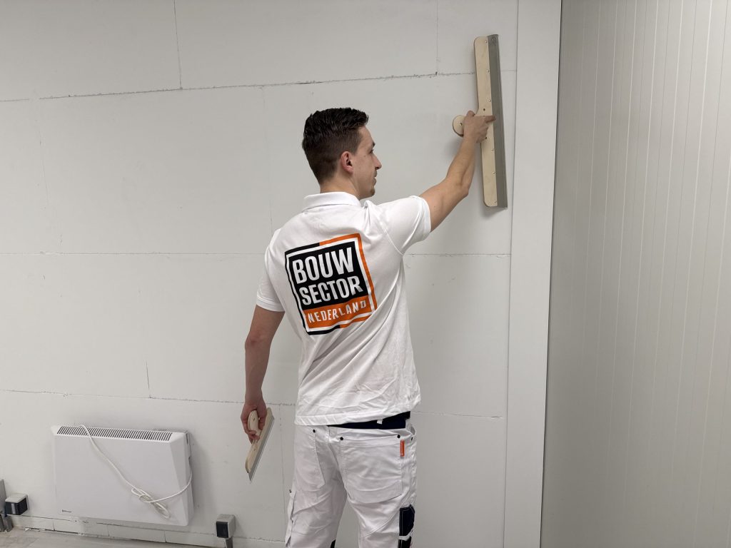 Stukadoor in wit poloshirt met het logo van Bouwsector Nederland trekt een wand vlak met een lange spaan
