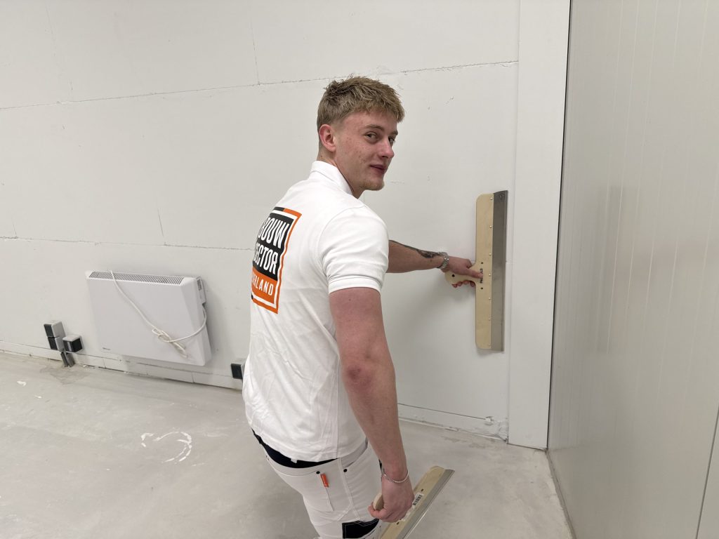 Stukadoor kijkt lachend om terwijl hij met een spaan een wand vlak bij de vloer glad zet