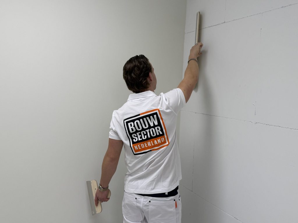 Stukadoor met Bouwsector-logo trekt de wand vlak bij de hoek van de kamer met een lange spaan